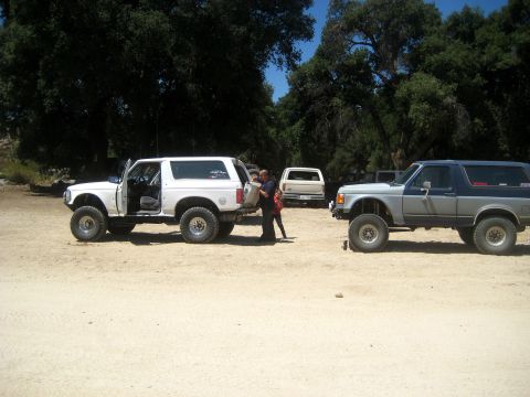 1996 Ford Bronco - Two Ford Broncos and a pickup truck are parked in a dirt lot with trees in the background.