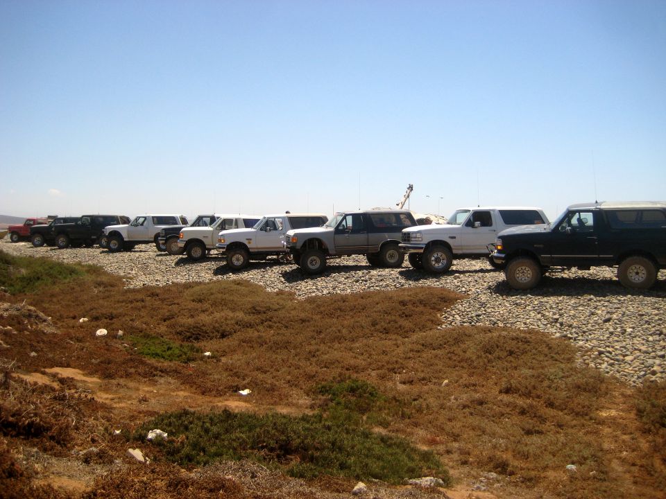 Line of Ford Broncos parked on a rocky shore under a clear blue sky