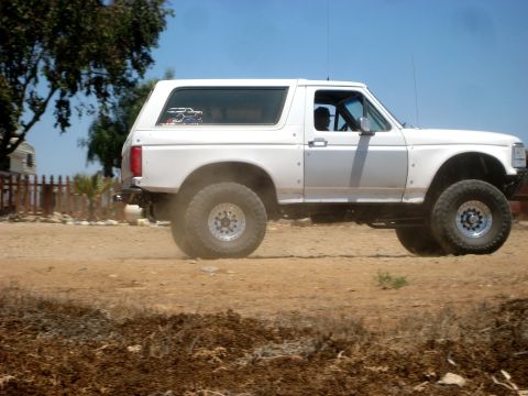 1996 Ford Bronco - White Ford Bronco with lifted suspension and off-road tires driving on a dirt road.