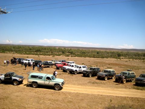 1996 Ford Bronco - A collection of older model SUVs and trucks are gathered on a dry, dusty road under a clear blue sky.