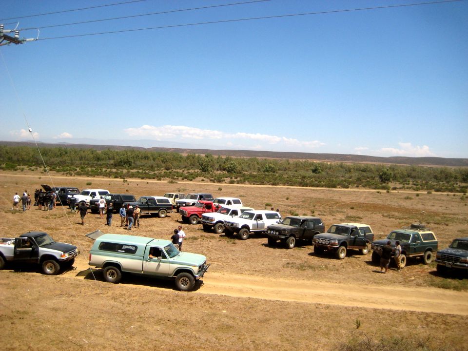 Several SUVs and trucks parked on a dirt road with a wooded area and hills in the background