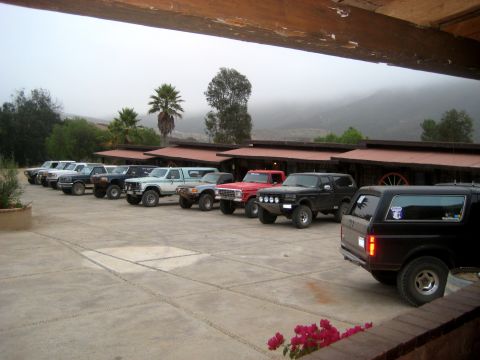 1996 Ford Bronco - Several Ford Broncos and trucks are parked in a lot outside a building with a mountain range in the background.