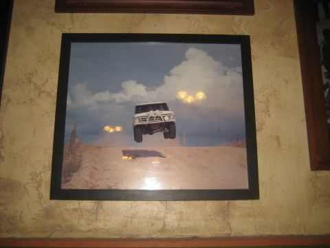 1996 Ford Bronco - This white SUV is captured mid-air, soaring over a dusty desert road under a cloudy sky.