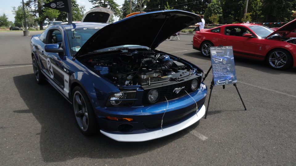 Blue Ford Mustang with hood open, red Ford Mustang in background. Display board next to blue car