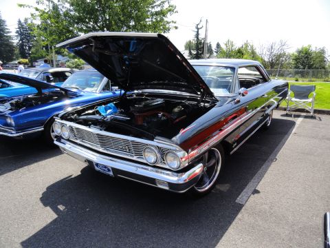 1964 Ford Fairlane - A black 1964 Ford Fairlane is displayed with its hood open, revealing the engine bay. Another blue car is visible to the left.