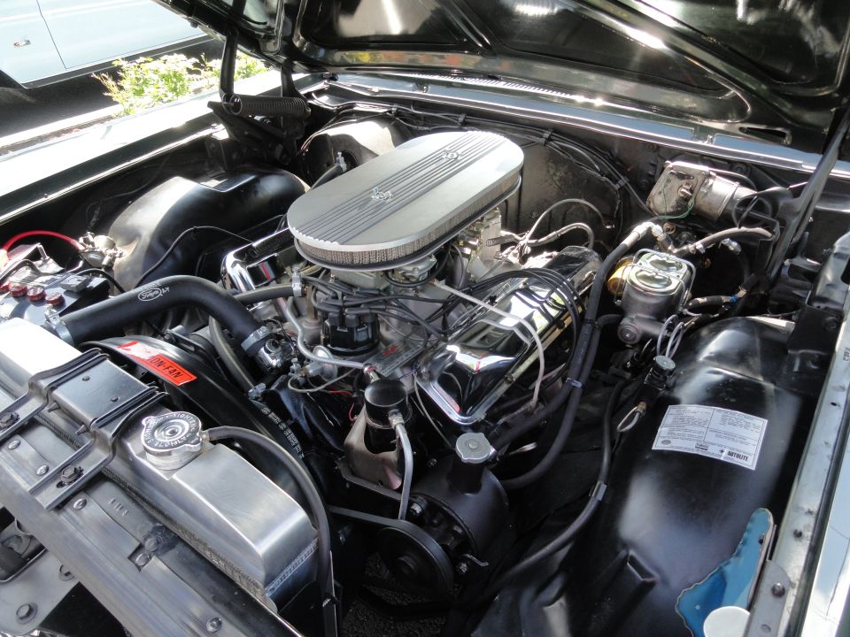 Engine bay of a black 1964 Ford Fairlane, showing a V8 engine with chrome valve covers and an oval air cleaner
