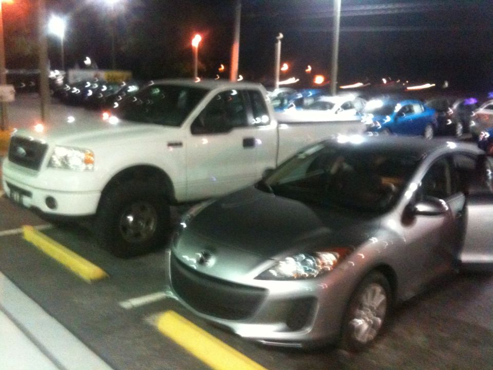 Silver Mazda sedan parked next to a white Ford pickup truck at night. The sedan's driver door is open