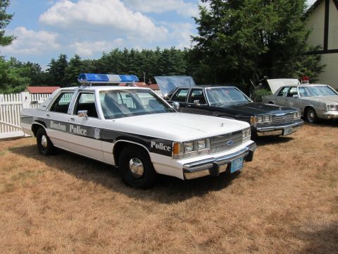 1991 Ford LTD Crown Victoria - black-and-white Boston Police Ford sedan with a blue light bar parked on grass beside other vintage cruisers.