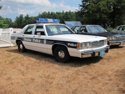 1991 Ford LTD Crown Victoria - black-and-white Boston Police Ford sedan with a blue roof light bar parked among other cruisers on grass.