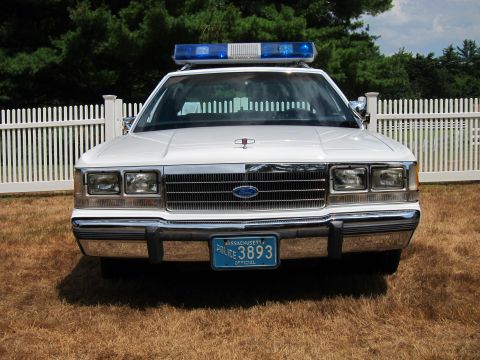 1991 Ford LTD Crown Victoria - Head-on view of a white police cruiser with a blue light bar, twin headlights and a Massachusetts 'Police 3893' plate on grass.