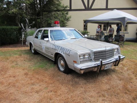 1991 Ford LTD Crown Victoria - silver full-size State Police sedan with a red roof light parked on grass near a tent and building.