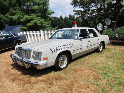 1991 Ford LTD Crown Victoria - tan Rhode Island State Police full-size sedan with a red roof light and satellite dishes behind it, parked on grass.