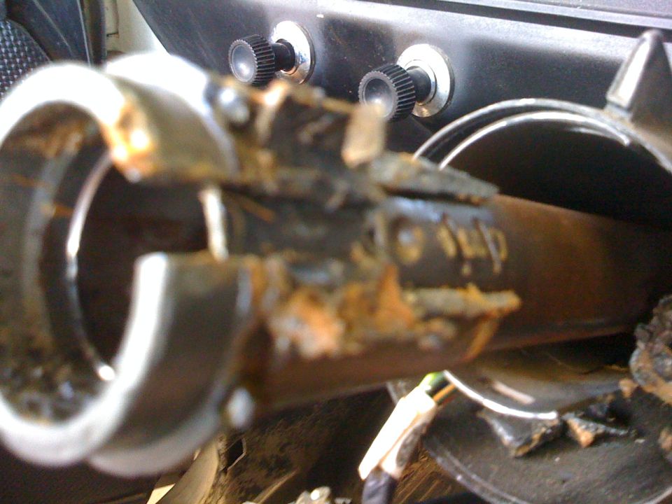Close-up of a rusted, broken steering column component with two knobs in the background