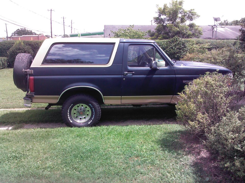 1994 Ford Bronco, dark blue with tan stripe, spare tire on rear door, parked on grass