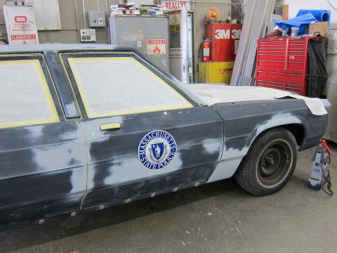 1991 Ford LTD Crown Victoria - A dark sedan, possibly a Ford Crown Victoria, is shown in a workshop with its body prepped for paint, a Massachusetts State Police emblem.