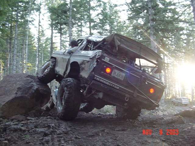 Maroon trail truck climbing a large rock at steep angle in the woods, driver visible, sun flare behind