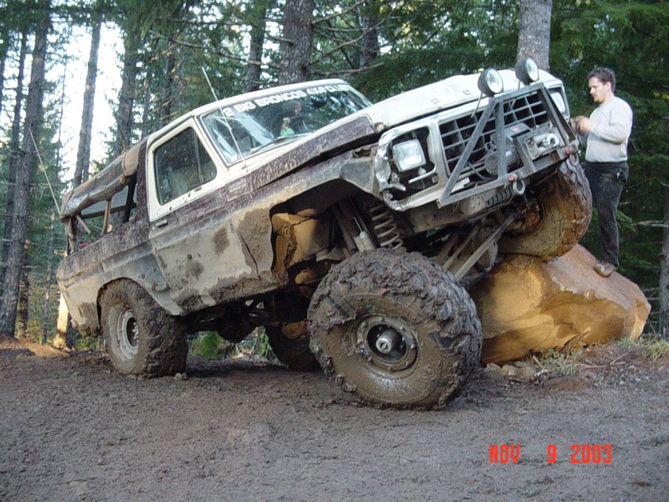 Muddy white-and-maroon trail truck with light bar perched on a large rock, man standing at right