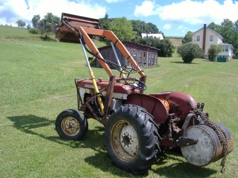 1988 Ford Bronco - A red tractor with a front loader attachment sits in a grassy field next to farm buildings.