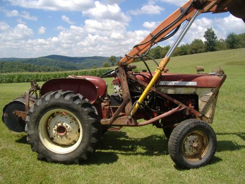 1988 Ford Bronco - A red tractor with a front loader attachment is parked in a grassy field with a cornfield and rolling hills in the background.