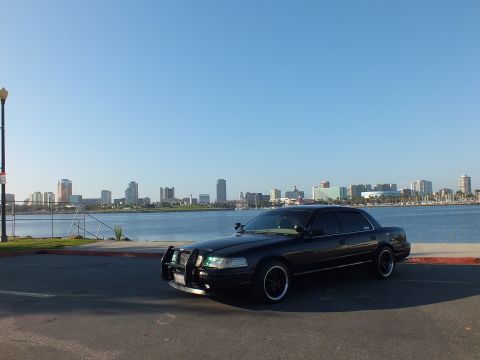 2003 Ford Crown Victoria - Dark police-style sedan faces a harbor skyline with headlights lit at dusk