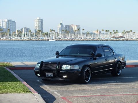 2003 Ford Crown Victoria - Dark sedan with brush guard glows headlights along the harbor curb with skyline