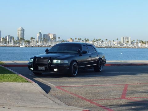 2003 Ford Crown Victoria - Blacked-out police-style sedan faces the bay with headlights on at the waterfront