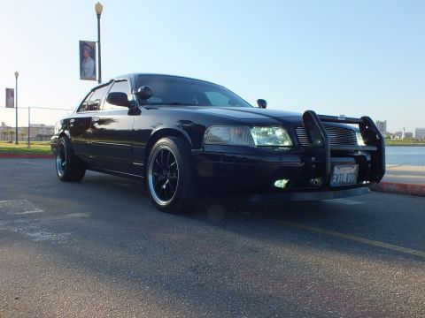 2003 Ford Crown Victoria - Murdered-out sedan with push bar and black wheels staged near waterfront banner poles