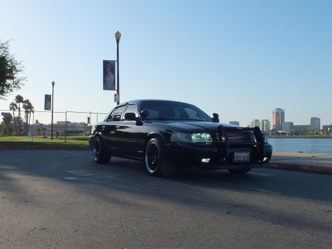 2003 Ford Crown Victoria - Lowered blacked-out sedan parked at an angle with city towers and palm trees in the distance
