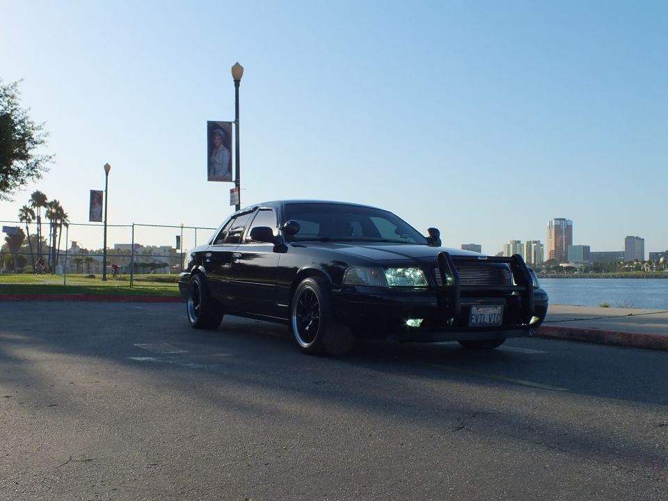 Black Ford Crown Victoria sedan with push bar in parking lot, skyline and banner poles behind