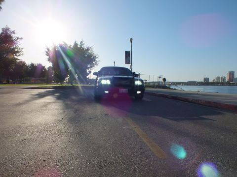 2003 Ford Crown Victoria - Dark sedan driving toward the camera with lights on, heavy lens flare from low sun over the water