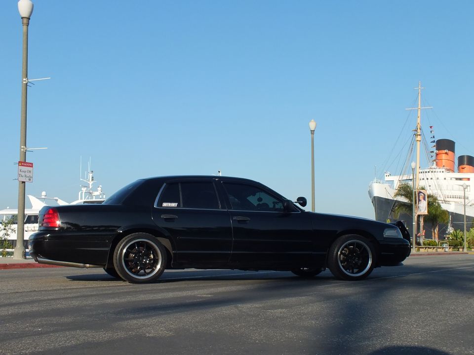 Side profile of black Ford Crown Victoria sedan on black wheels, Queen Mary ship behind