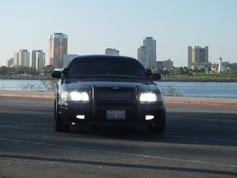 2003 Ford Crown Victoria - Blacked-out sedan facing camera with lights on and EVIL VIC plate, downtown towers behind