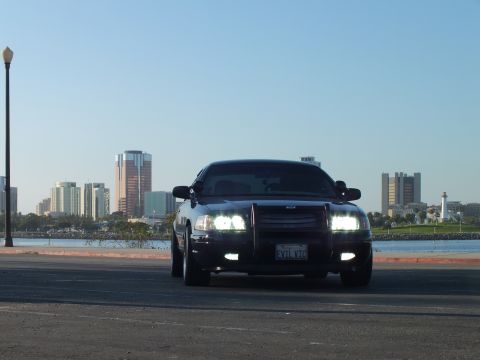 2003 Ford Crown Victoria - Lowered blacked-out sedan facing camera with push bar and lights on, downtown towers beyond