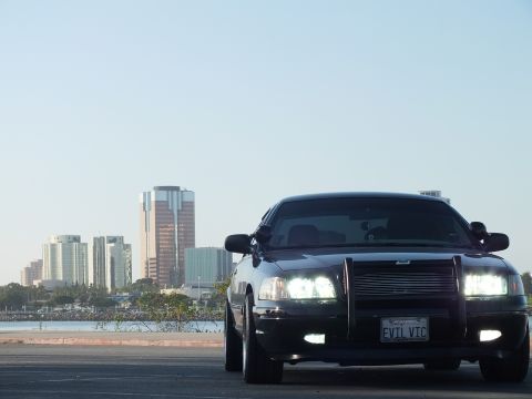 2003 Ford Crown Victoria - Murdered-out sedan angled toward camera with push bar and headlights on, skyline backdrop