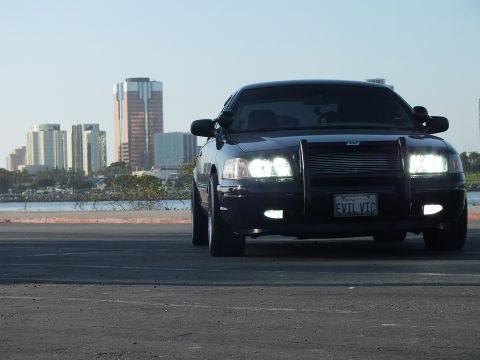 2003 Ford Crown Victoria - Front view of a blacked-out sedan with push bar and lights on against the downtown skyline