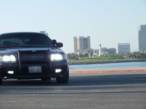 2003 Ford Crown Victoria - Murdered-out sedan angled toward camera with push bar, lighthouse and skyline across the water