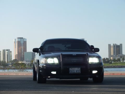 2003 Ford Crown Victoria - Front view of a lowered blacked-out sedan with push bar and lights on, skyline across the water