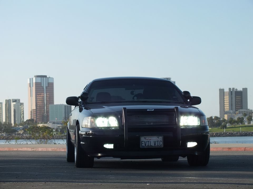 Black Ford Crown Victoria with grille guard and lit headlights, EVIL VIC plate, harbor behind