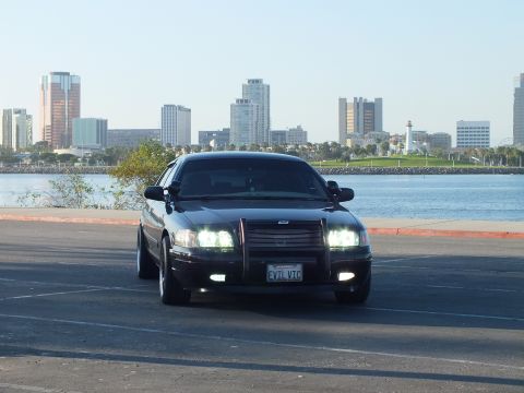 2003 Ford Crown Victoria - Murdered-out sedan facing camera with push bar and headlights on, lighthouse and towers behind