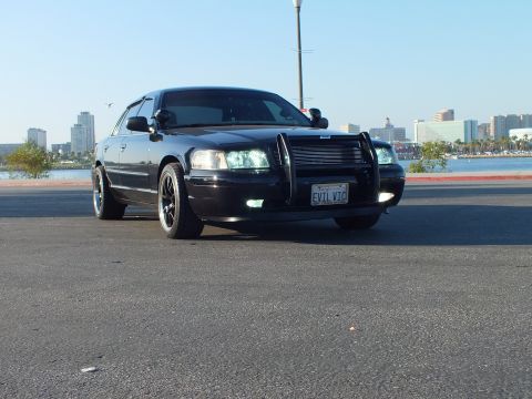 2003 Ford Crown Victoria - Front three-quarter view of a blacked-out Crown Victoria, grille guard fitted, city skyline and bay behind.