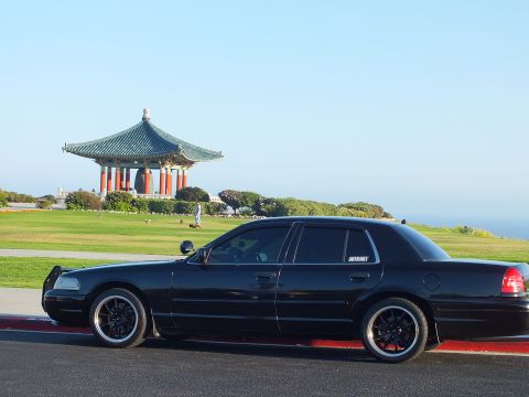 2003 Ford Crown Victoria - Side profile of a lowered black sedan with gloss-black multi-spoke wheels; pagoda pavilion and sea in the distance.