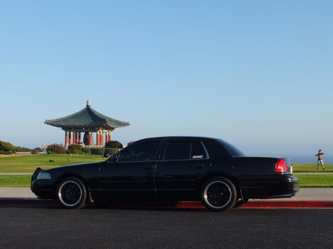 2003 Ford Crown Victoria - Profile shot of a black four-door on black rims along a curb, pagoda and coastline behind.
