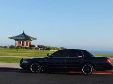 2003 Ford Crown Victoria - Rear-quarter view of a lowered black sedan on dark alloys, pagoda pavilion on grass behind.