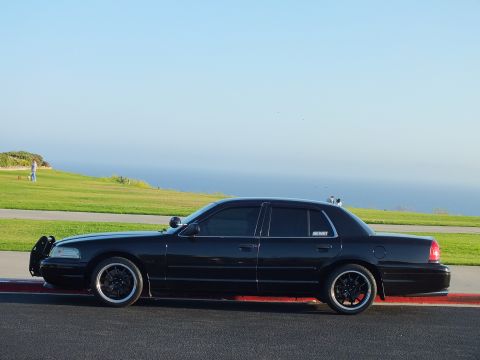 2003 Ford Crown Victoria - Rear-quarter angle of a blacked-out sedan with grille guard and dark wheels above a coastal bluff.
