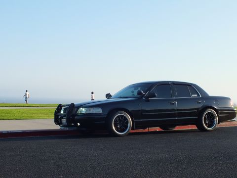 2003 Ford Crown Victoria - Front-quarter view of a lowered black four-door fitted with a brush guard, pedestrians on the grass behind.