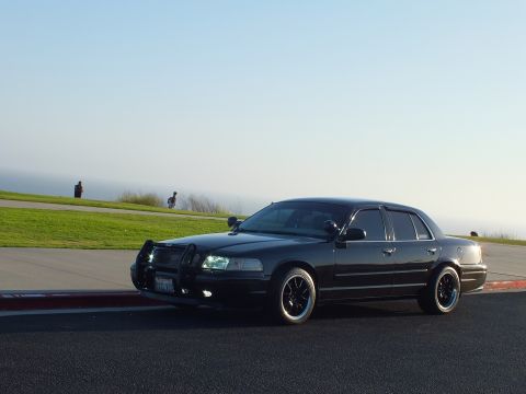 2003 Ford Crown Victoria - Front three-quarter of a black sedan with chrome push bar and headlights on, hazy coast behind.