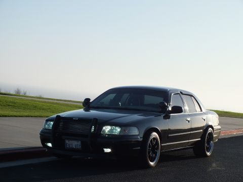 2003 Ford Crown Victoria - Front-quarter view of a blacked-out four-door, push bar and headlights lit, coastal grass behind.