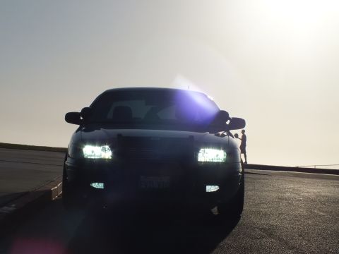 2003 Ford Crown Victoria - Front view of a black car with headlights on full, heavy lens flare from the sun overhead.