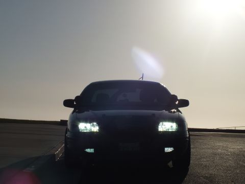 2003 Ford Crown Victoria - Head-on silhouette of a black four-door, headlights lit and antenna raised against a bright sky.