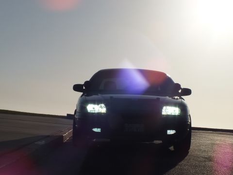 2003 Ford Crown Victoria - Frontal shot of a black car with lights on, strong sun flare washing across the windshield.